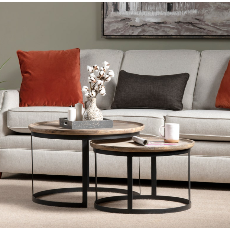 Living room setup with a beige couch adorned with two rust-colored pillows and a dark brown pillow. In front, two Fillmore Nesting Coffee Tables of different heights, each with a mango wood top and black metal base. Tables hold decorative items: a vase with cotton, a tray, and candles. Wall art is hung above the couch.
