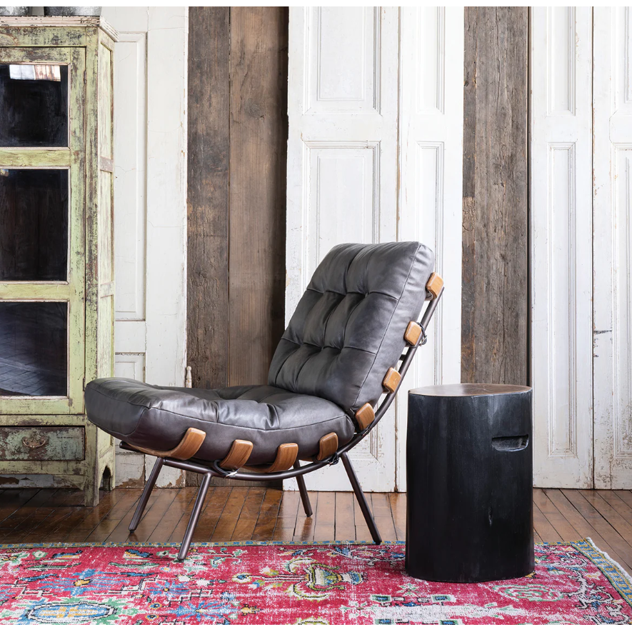 A modern room features the gray button-tufted leather Lance Lounge Accent Chair with wooden accents on a metal frame, placed next to a small black cylindrical side table. The backdrop consists of rustic wooden panels, and a colorfully patterned rug adds a pop of color to the mid-century style wooden floor.