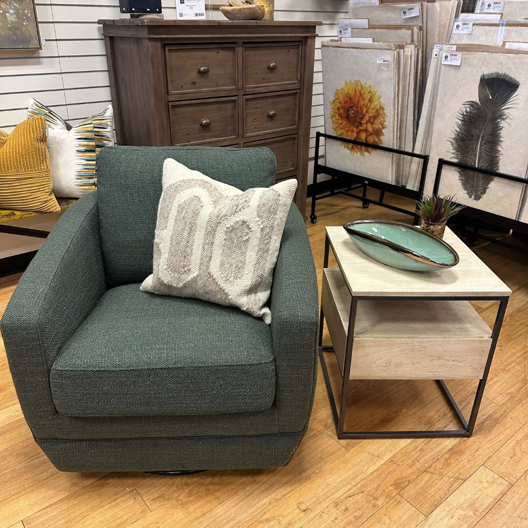 A cozy setup includes a dark green armchair with a white pillow. Beside it, the Halston Side Table in natural wood holds a ceramic dish and small plant. Framed artworks adorn the backdrop against a wooden cabinet on the wood floor.