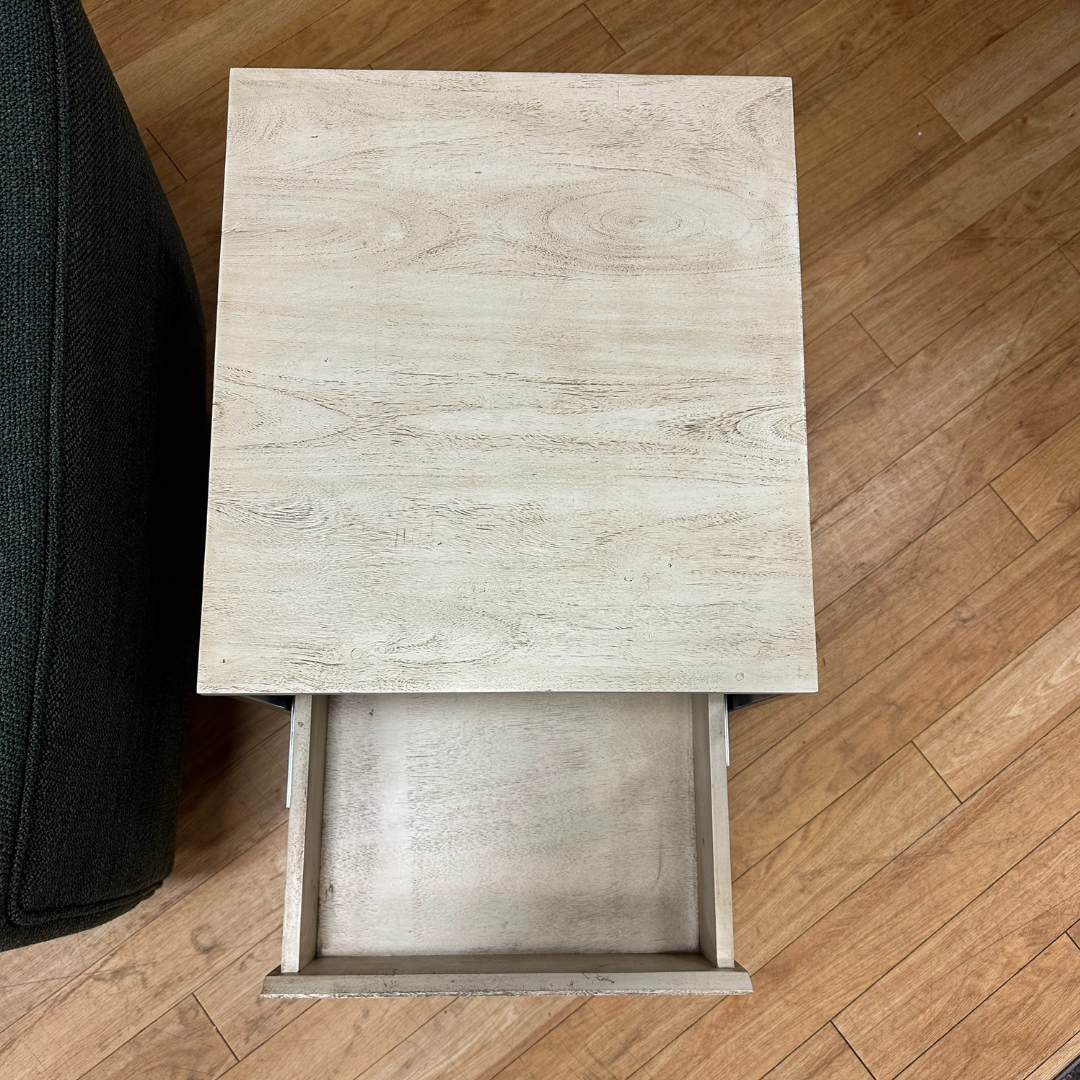 A Halston Side Table with a light natural wood finish is next to a dark textured sofa. Its open drawer shows an empty interior, while the unit rests on a wooden floor with distinct grain patterns, adding a modern touch to the room.