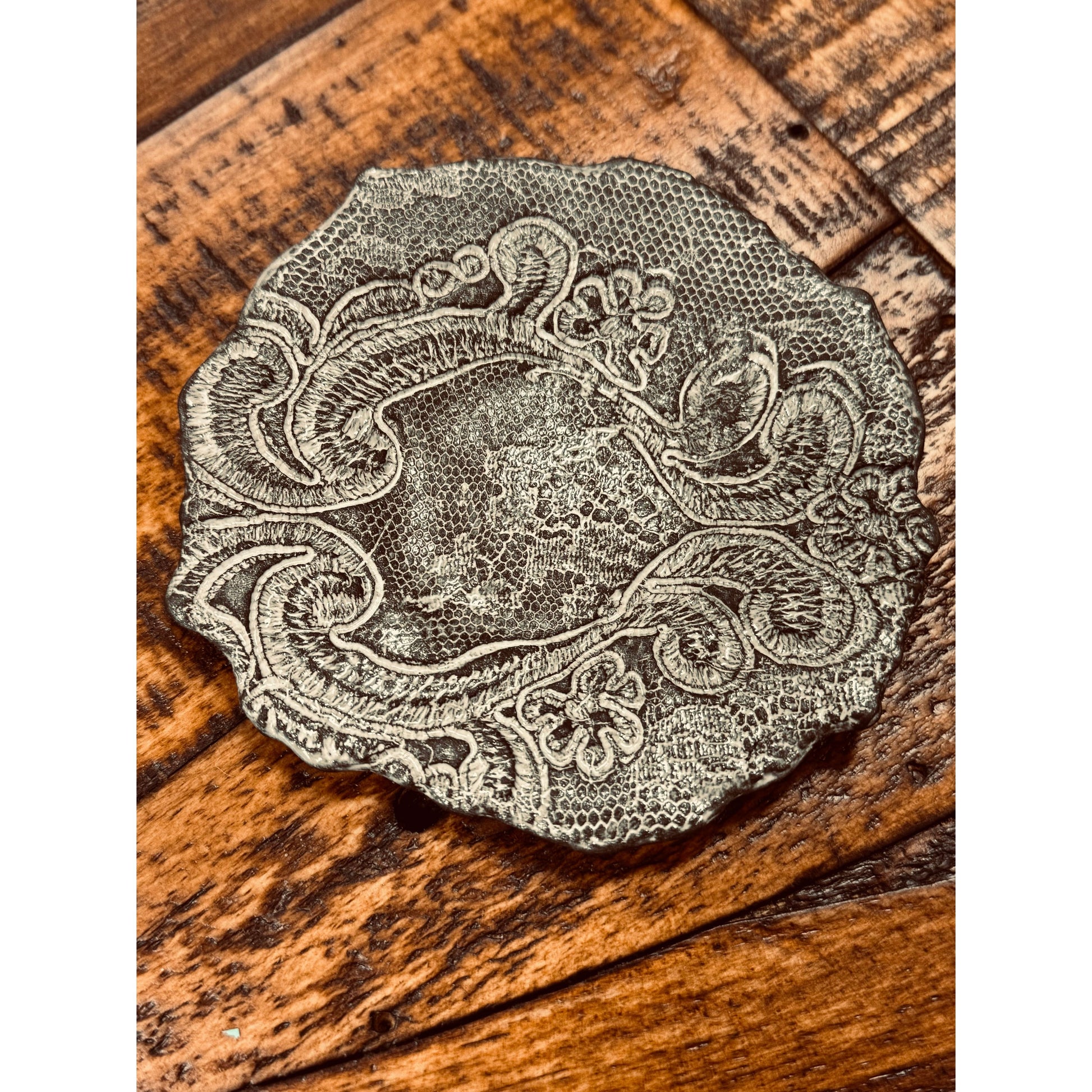 A round, intricately designed metal object lies on a wooden surface. The object features ornate, swirling patterns and floral motifs, reminiscent of a Casada Jewelry Dish. The wood beneath it has a rich, warm tone with visible grain and hints of wear, highlighting its rustic charm.