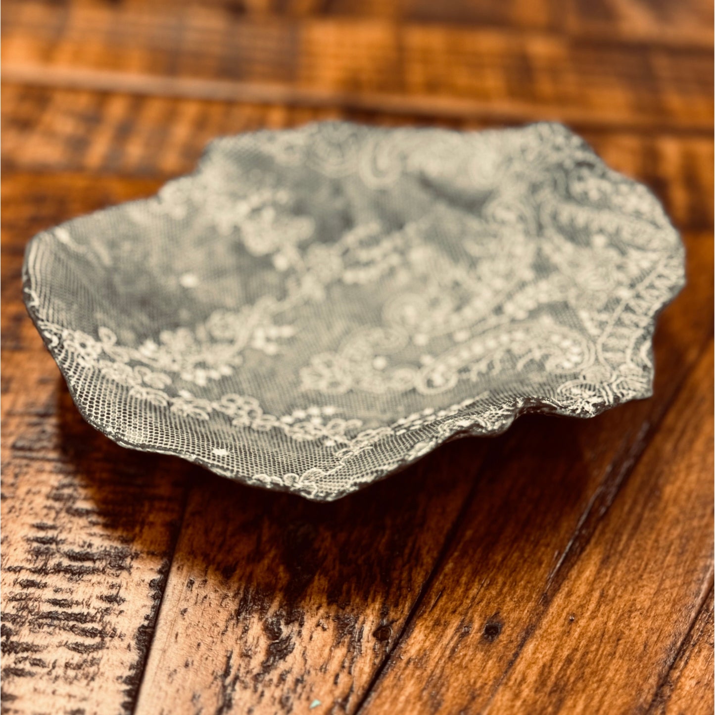 A delicate, intricately detailed doily lies on a wooden surface. The doily features a lace pattern with floral and swirling designs. Next to it, the Casada Jewelry Dish by a skilled ceramic artist complements the scene, with the wooden background showcasing visible grain and adding a rustic contrast.