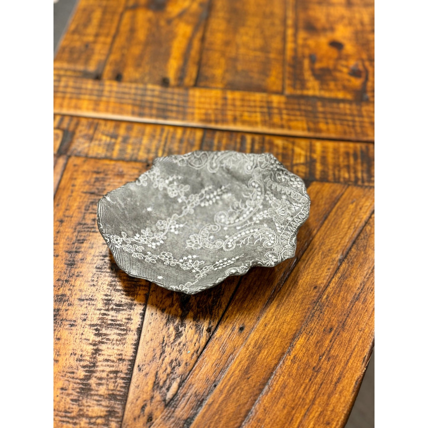 A small, intricately patterned black Casada Jewelry Dish by Valerie Casado ceramics rests on a rustic wooden surface. The dish's delicate design contrasts with the textured and slightly worn wood grain, adding to its vintage aesthetic. The wood shows various shades of brown, highlighting its natural imperfections and beauty.