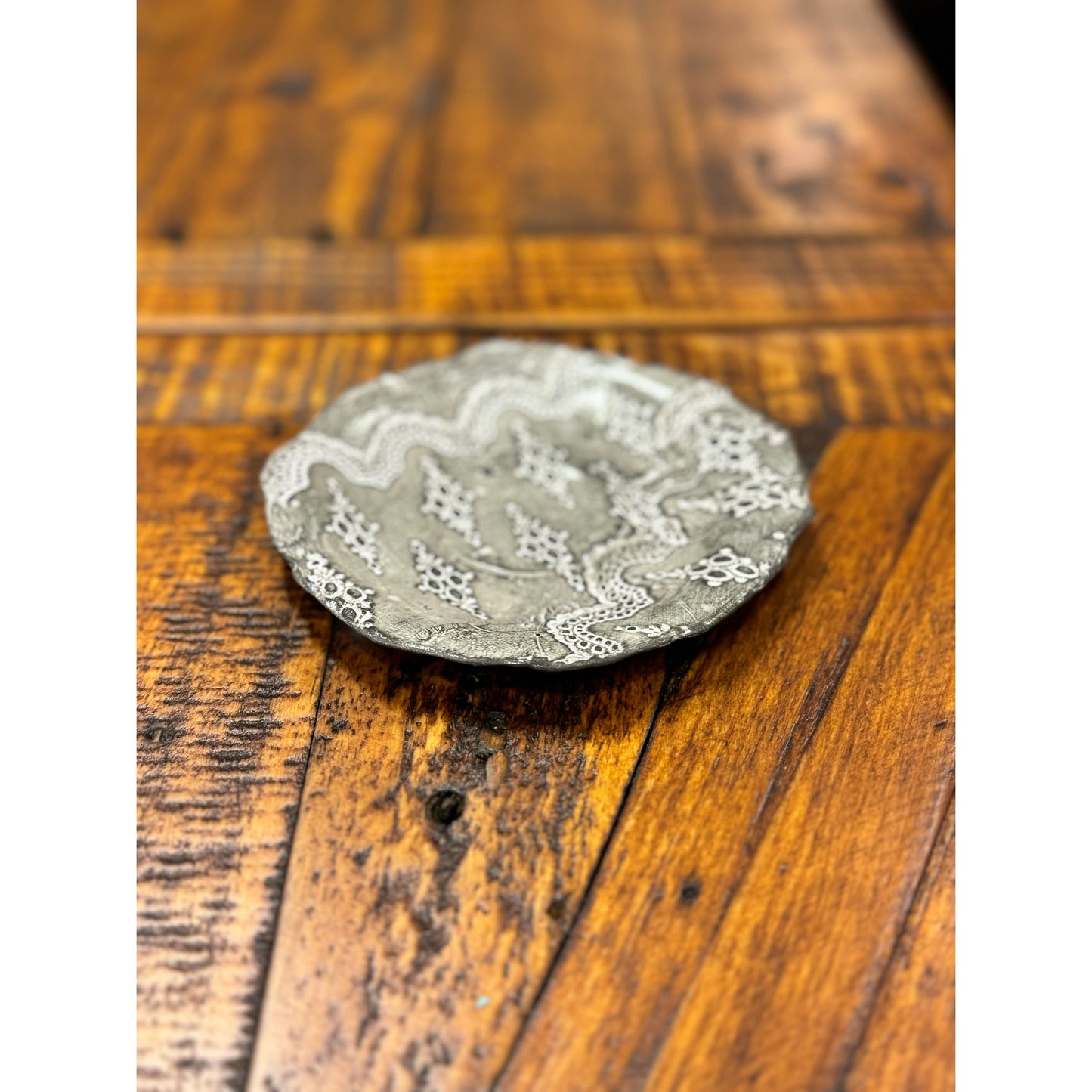 A round ceramic Casada Jewelry Dish with intricate white lace-like patterns, a fine example of Valerie Casado ceramics, is centered on a rustic wooden table. The table's surface shows rich textures and wood grain, featuring a mix of lighter and darker brown tones along with visible knots and plank lines.