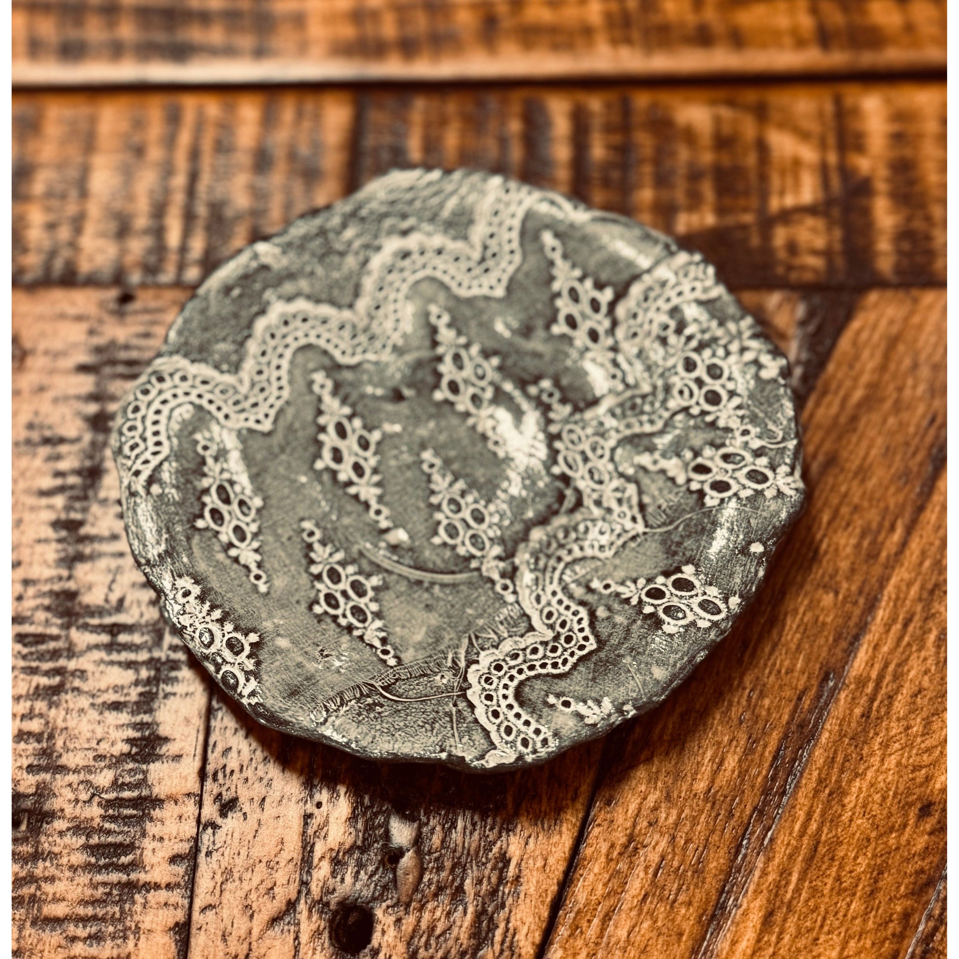 A round, textured clay piece with intricate lace-like patterns sits on a wooden surface. The patterns, reminiscent of delicate doilies, cover the gray, rustic clay, adding an elegant contrast. This Casada Jewelry Dish rests on a wooden surface showing varying shades of brown and visible grain.