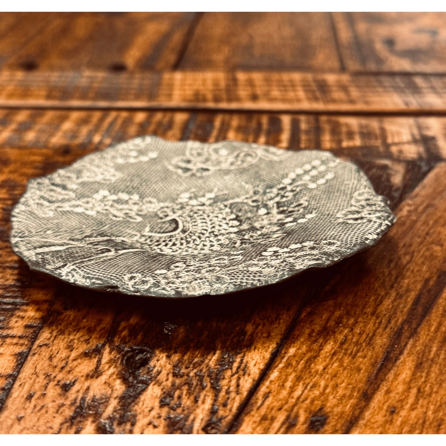 A close-up of a decorative Casada Jewelry Dish set on a wooden table. The dish features intricate floral and geometric patterns with a slightly scalloped edge. The wood surface shows a rich, textured grain and warm, varied tones, with the dish positioned slightly off-center like unique ceramic art by Valerie Casado ceramics.