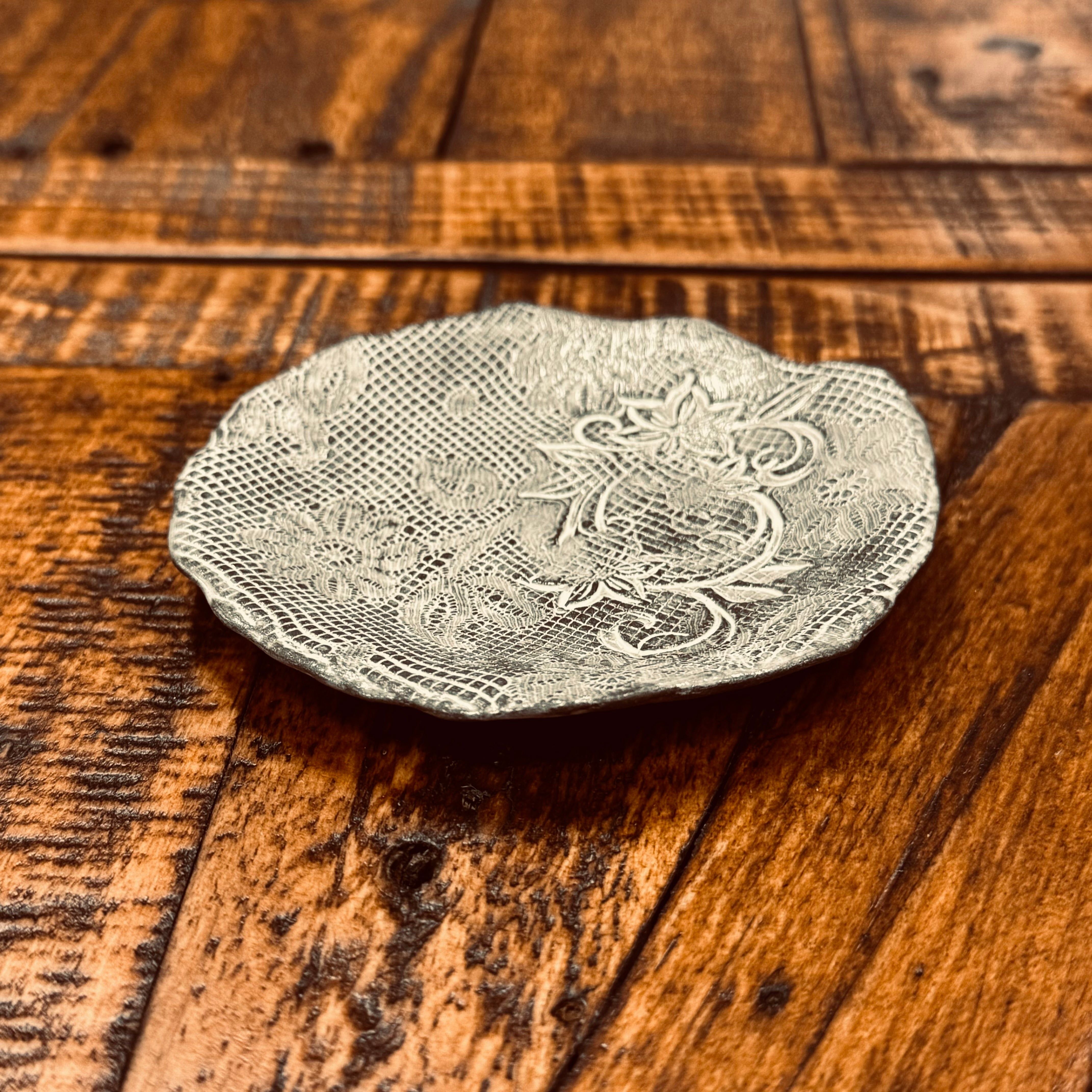 A small, intricately designed Casada Jewelry Dish lies on a wooden surface. The dish features elaborate, textured patterns and floral motifs reminiscent of handcrafted pottery by Valerie Casado ceramics. The warm tones of the wooden surface contrast with the silvery hue of the dish.