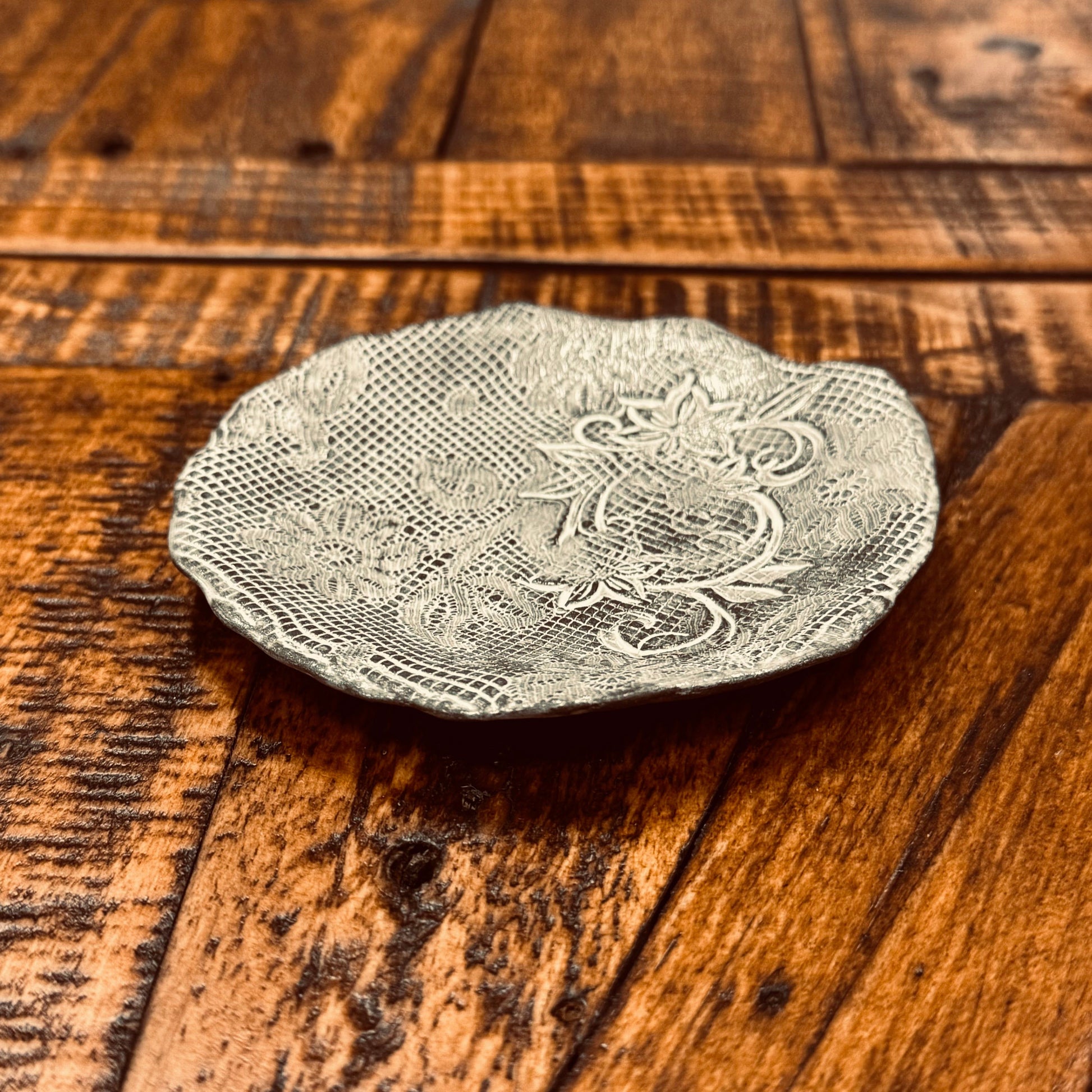 A small, intricately designed Casada Jewelry Dish lies on a wooden surface. The dish features elaborate, textured patterns and floral motifs reminiscent of handcrafted pottery by Valerie Casado ceramics. The warm tones of the wooden surface contrast with the silvery hue of the dish.