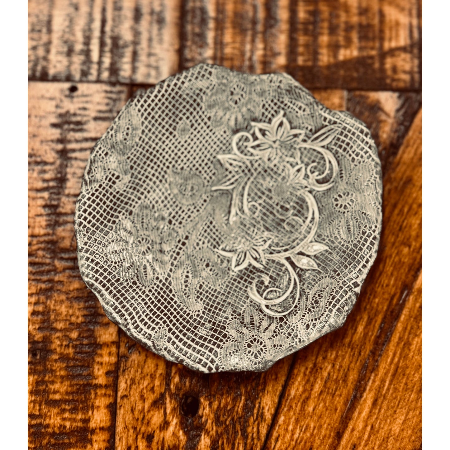 An intricately designed silver Casada Jewelry Dish sits on a rustic wooden surface. The dish features delicate floral and lace-like patterns with ornate scrollwork, creating a textured and elegant appearance reminiscent of Valerie Casado ceramics. The wood background has varied shades of brown, highlighting the dish's detail.