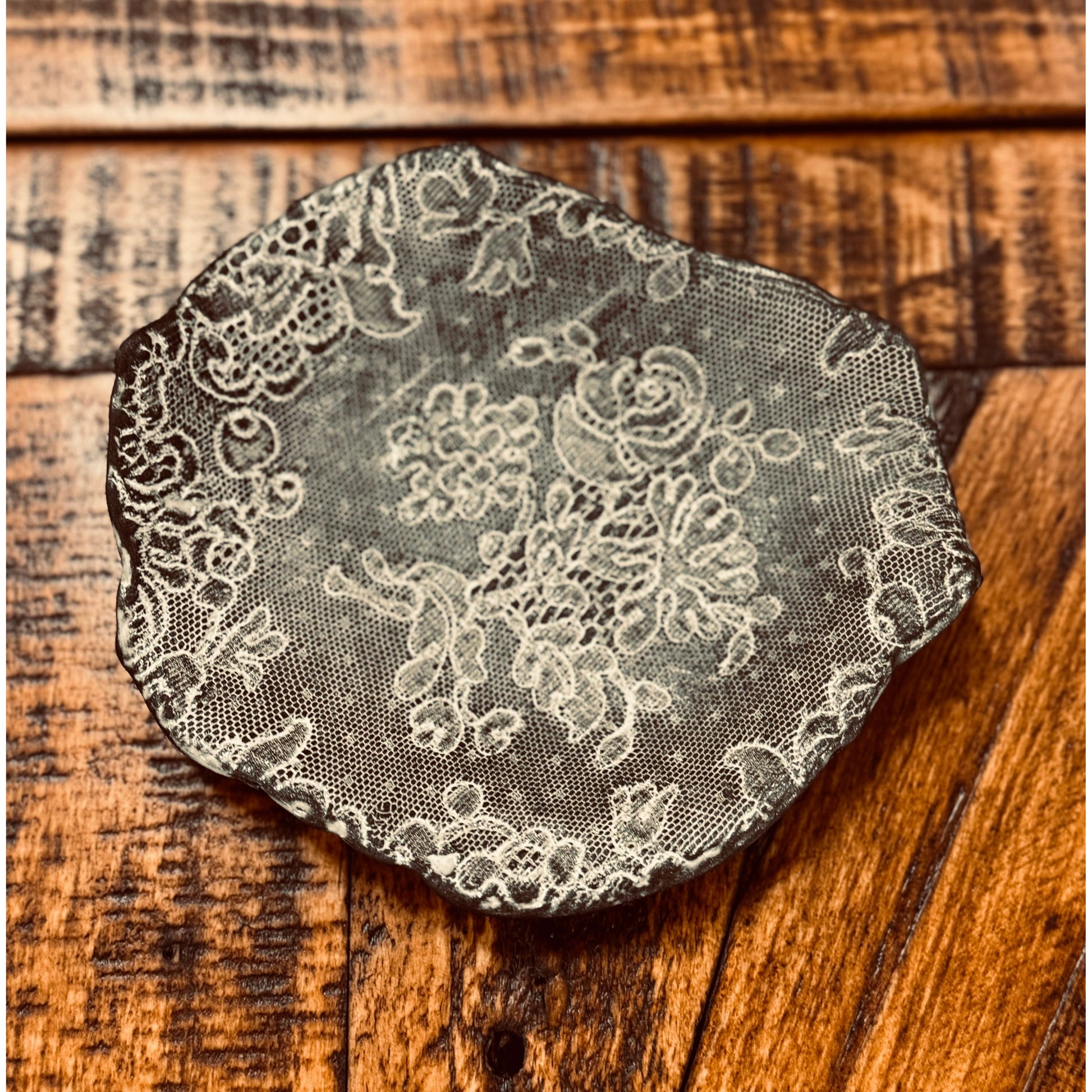 A round, black ceramic dish with textured white lace patterns, created by Casada Jewelry Dish, is placed on a rustic wooden surface. The handcrafted pottery features intricate floral designs and uneven edges, giving it an artisanal appearance. The wood's rich grain and worn texture complement the delicate details of the dish.