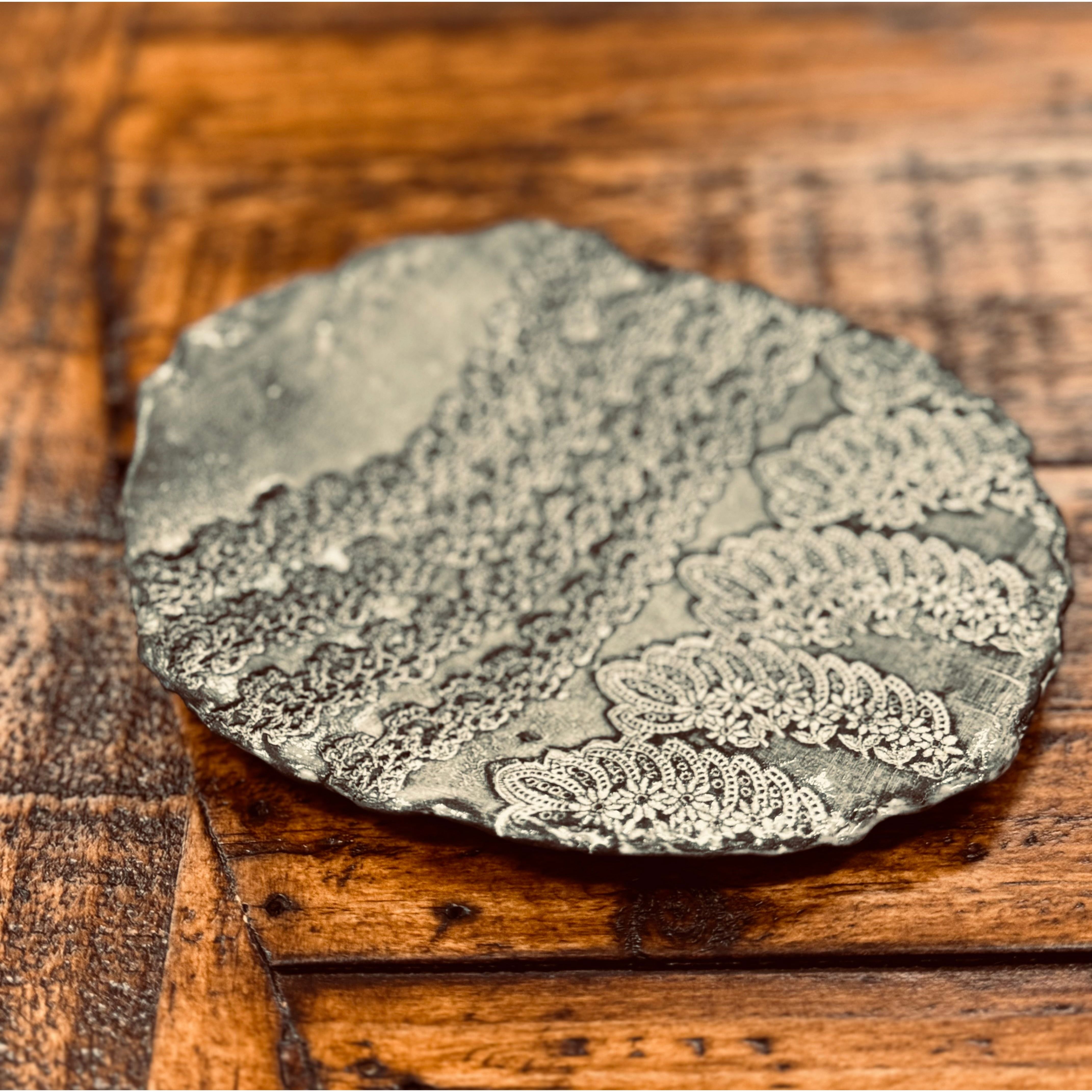 A Casada Jewelry Dish featuring intricate, ornate patterns resembling lacework. The patterns consist of wavy and circular designs, with some resembling leaf shapes. Reminiscent of Valerie Casado ceramics, it sits on a dark, textured wooden surface, adding a rustic contrast to the detailed craftsmanship.