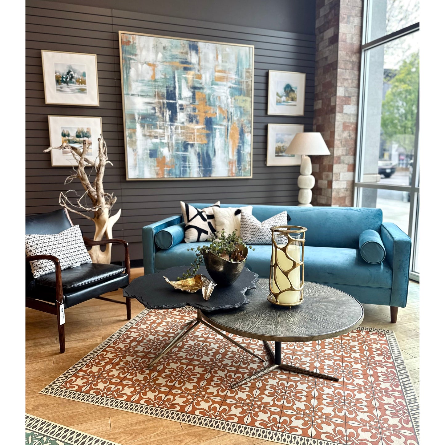 A stylish living room featuring a Bench Velvet Blue Sofa, a black leather chair, a circular metal coffee table, and a vibrant abstract painting above the couch. The room has wood paneled walls and large windows.