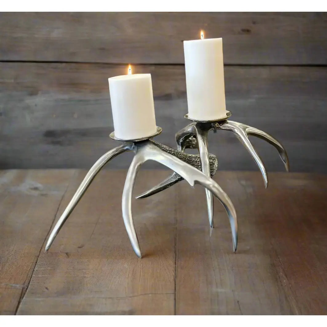 The Antique Silver Antler Candleholder, made from recycled aluminum, gracefully holds two white pillar candles. Its authentic silver finish mimics the natural curves of deer antlers. Placed on circular bases against a neutral gray background, these candlesticks highlight their brilliant metallic shine.