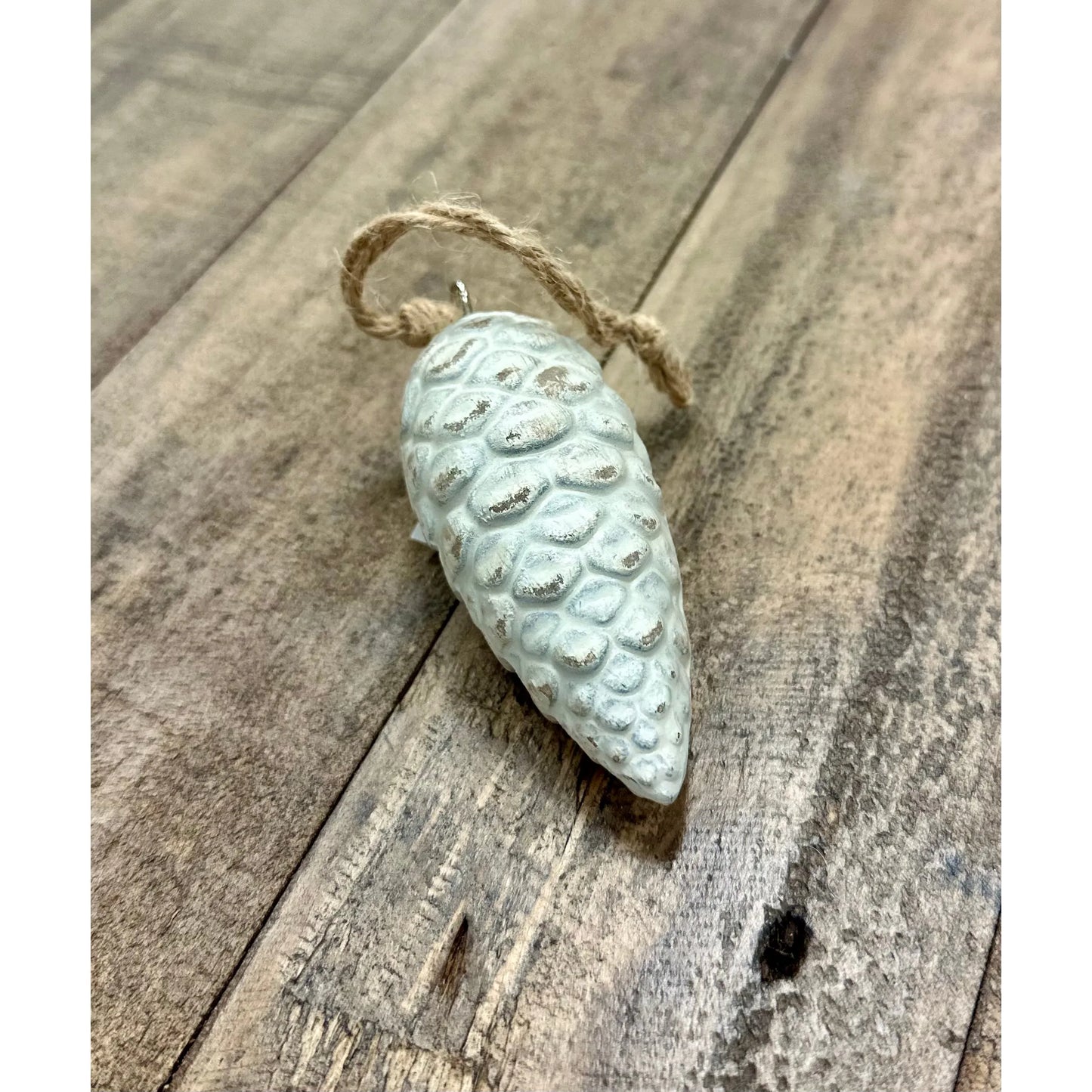 The *Wood Pinecone Ornament, Small* is displayed on a wooden surface, showcasing its white pinecone shape and rustic finish. It includes a natural twine loop for hanging and measures 3 inches. The wood grain of the background enhances its holiday decor appeal.