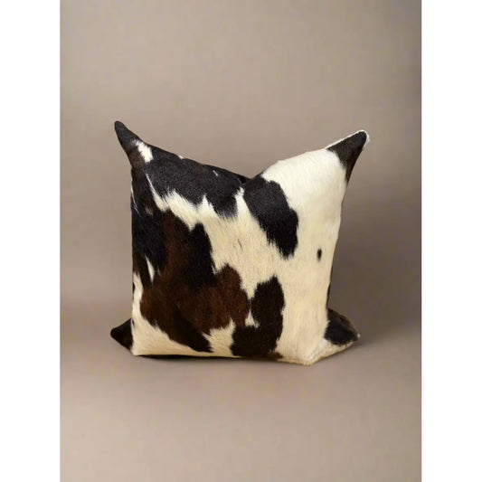 The Hair on Hide Throw Pillow showcases an intricate cowhide pattern with unique black and white patches on a light brown background. Made from natural materials, it stands out against a simple, neutral setting, emphasizing its texture and striking design.