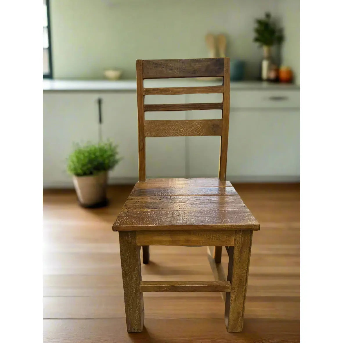 In a sunlit kitchen, a Wood Dining Chair, showcasing a rustic handcrafted solid wood design with a slatted backrest, rests elegantly on the wooden floor. The blurred background reveals rustic charm, featuring a small plant to the left and kitchen items to the right on the counter.
