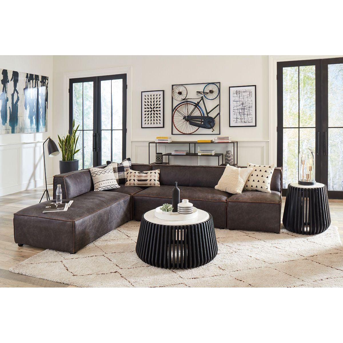 A modern living room featuring an Antique Ebony Leather Sofa with matching high density foam cushions, two unique black coffee tables, a beige rug, and wall art depicting abstract and bicycle themes. Natural light streams in from large windows.