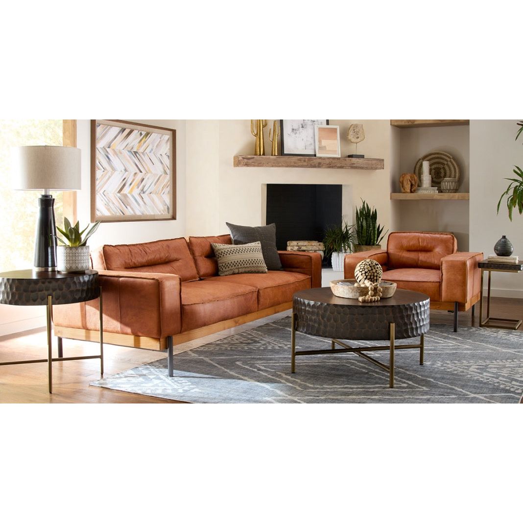 A cozy living room featuring a tan leather sofa set including a three-seater and two Cocoa Leather Accent Chairs with high density foam cushions around a black round coffee table, on a gray rug. A mantelpiece with decorative items, abstract wall art, and a lamp add warmth to the space.