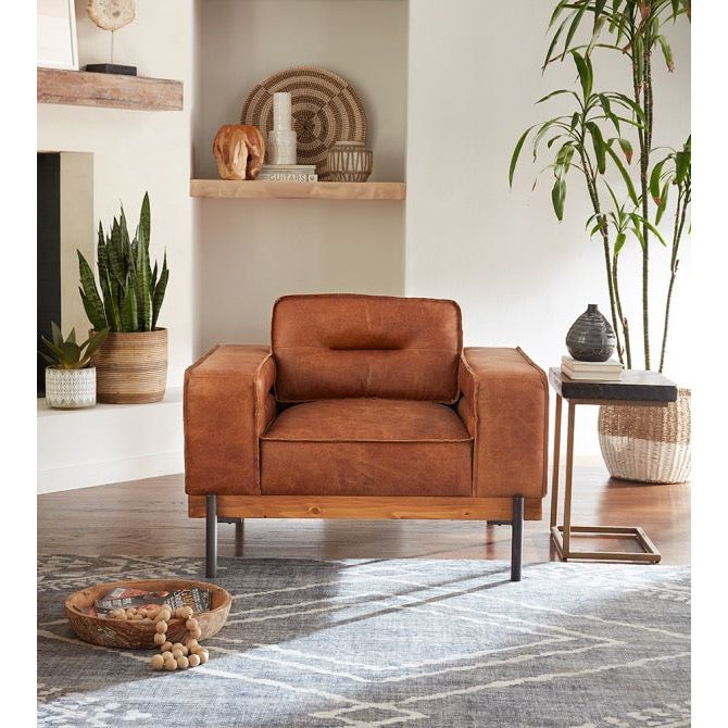 A stylish living room featuring a Cocoa leather accent chair with wooden legs on a patterned gray rug. To the left, a shelf with wicker baskets and a decorative item, and to the right, a tall potted plant beside a small table with a lamp.