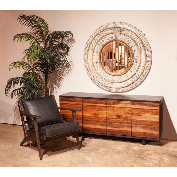 Sentence with Product Name: A stylish interior corner with a sleek wooden sideboard featuring varied wood patterns, placed beneath a circular mirror with a sunburst frame. A comfortable Dalton Rustic Leather Armchair and a lush green potted plant are to the side, enhancing the warm ambiance.