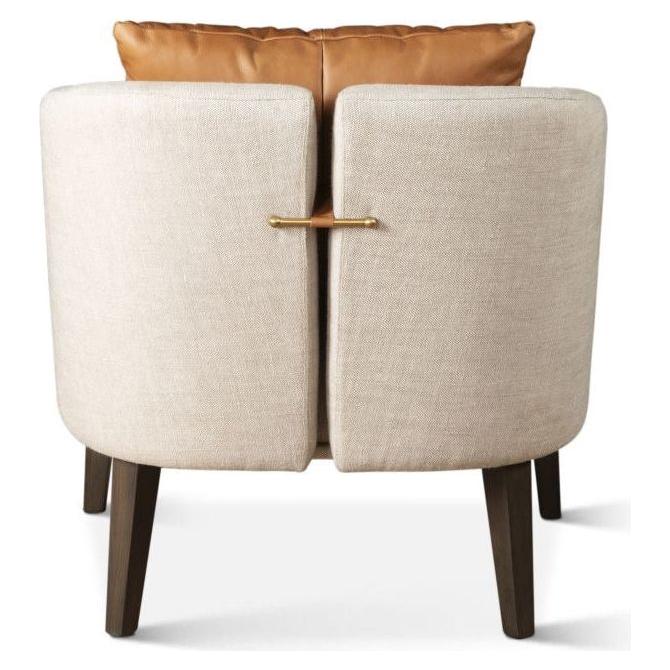 The Serenity Accent Chair, shown from behind, boasts a beige linen upholstery with a curved back. It features a decorative brass bar connecting two back panels and stands on four dark wooden legs. A brown leather cushion at the top adds stylish contrast to this luxurious design.