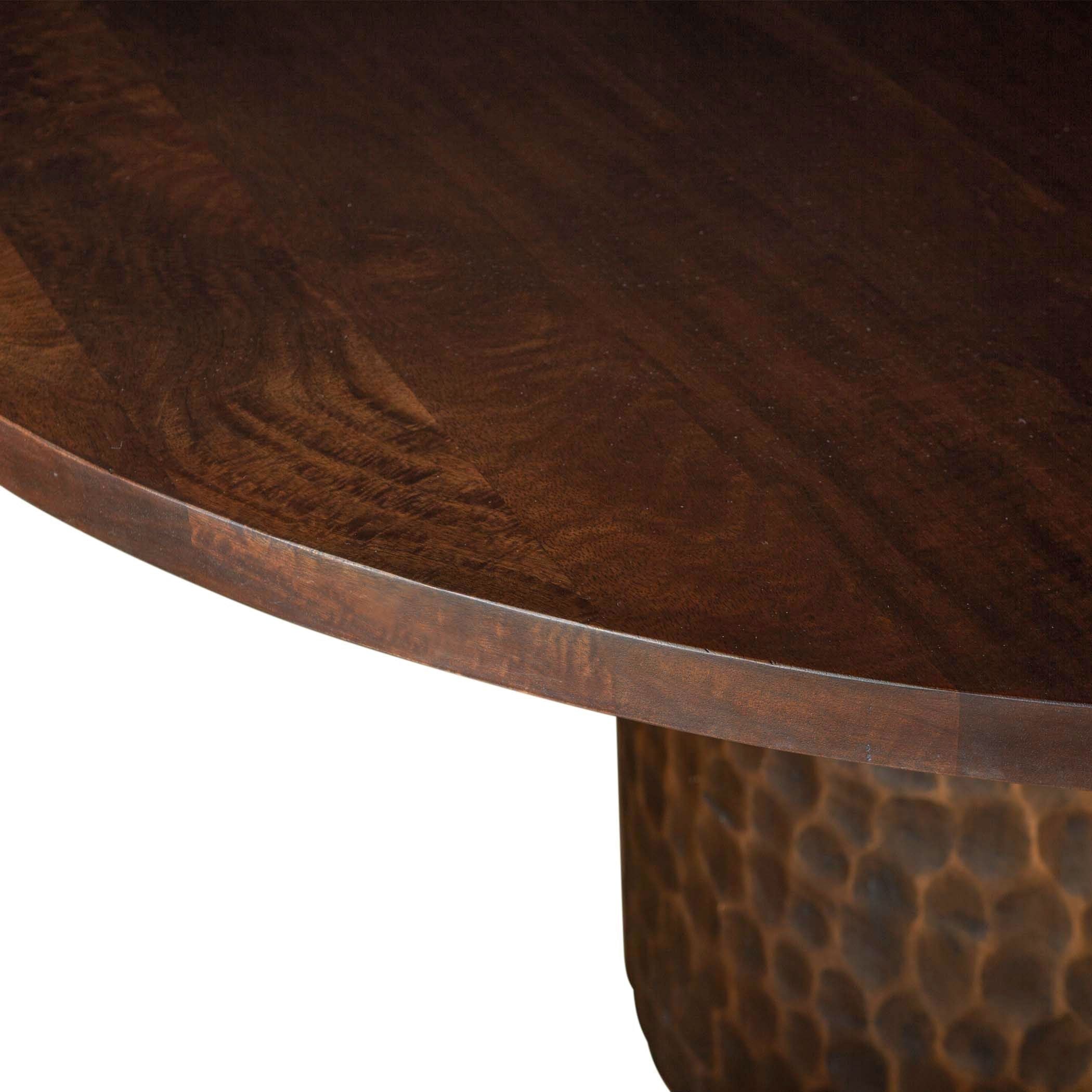 The Cruz 60" Round Dining Table features a close-up of a rich, dark brown mango wood tabletop with visible grain patterns atop a cylindrical base that has a hammered metal appearance, offering a sleek contrast between smooth wood and rugged metal.