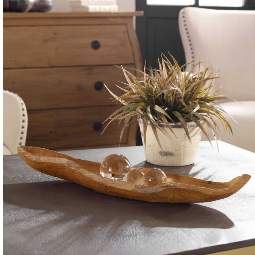 A Luvira Teak Leaf Bowl with three glass spheres rests on a gray table. A spiky-leafed potted plant in a light-colored pot adds charm. In the background, a wooden chest of drawers and a beige upholstered chair catch light near a large window.