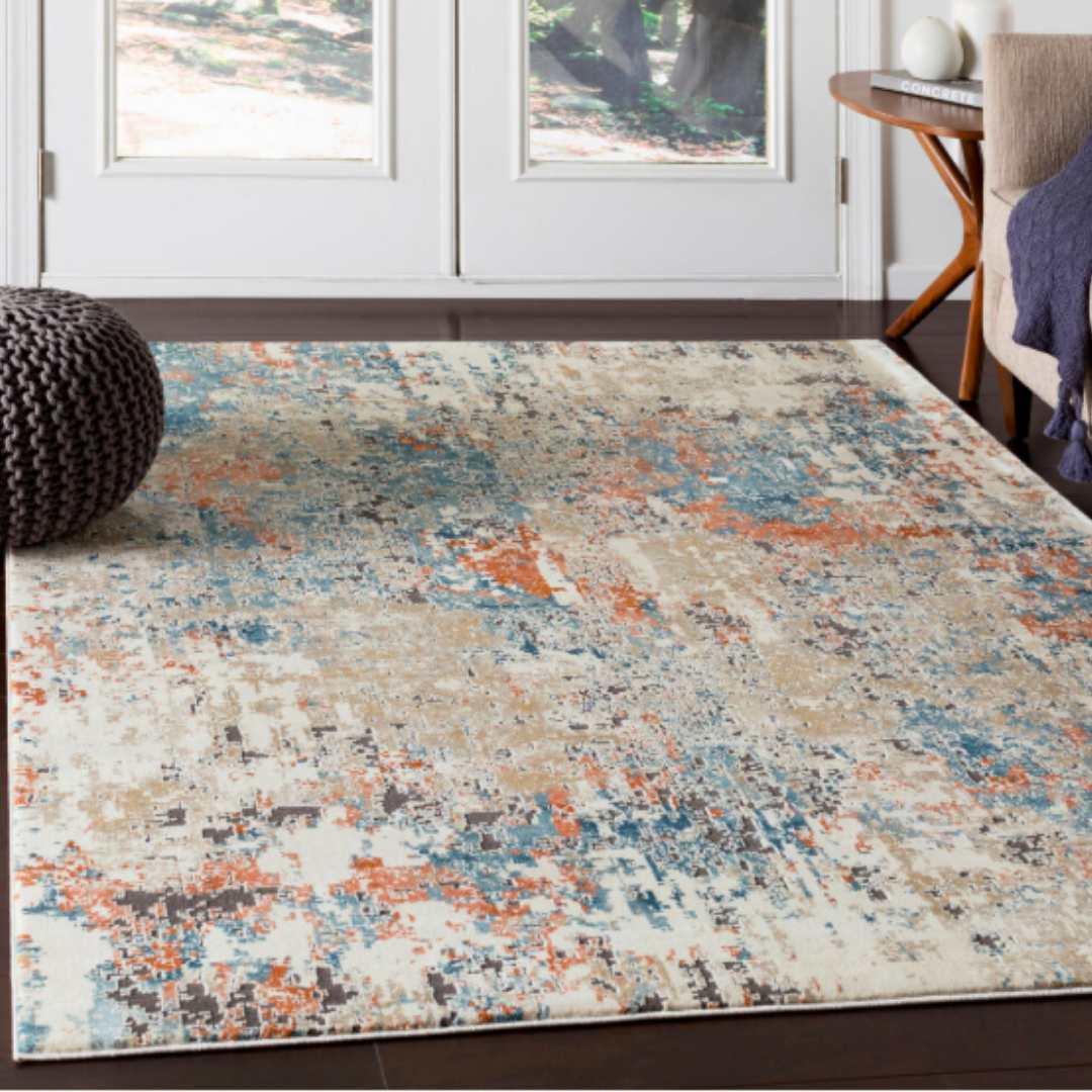 A modern living room features the Aurora Machine Woven Rug in polypropylene, displaying beige, orange, blue, and gray tones—ideal for transitional spaces. It pairs well with a round knit pouf, white table with books, and garden-facing glass doors.