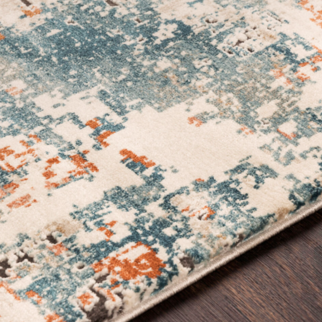A close-up of the Aurora Machine Woven Rug shows an abstract pattern in teal blue, orange, and beige. Made from polypropylene, it suits transitional interiors and is displayed on a textured dark wood floor with color variations.