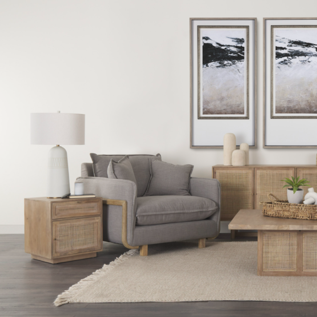 A cozy living room with a Veyra Plush Upholstered Armchair in Earthy Grey, a light wood side table topped with a white lamp, and a stylish coffee table. Dark wood flooring with a beige rug unifies the space, adorned by abstract art, plants, and sculptural accents.