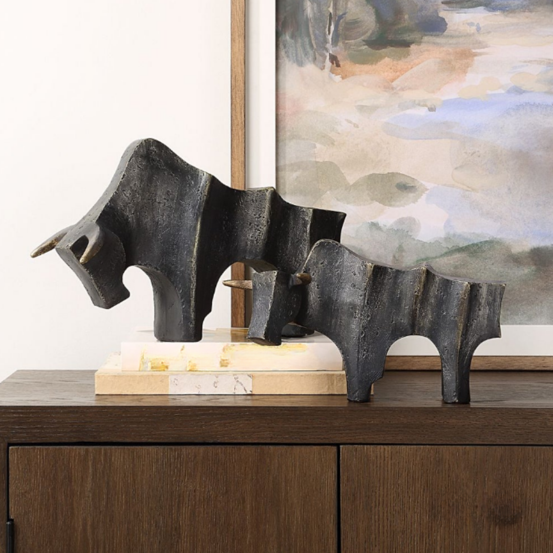 Two Noble Bull Statues with gold accents stand on stacked white and gold books atop a wooden cabinet. Behind them, a framed landscape painting with muted colors decorates the white wall.