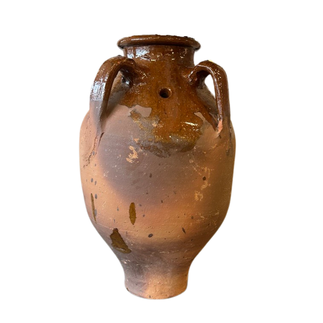 The Olive Jar is a rustic, weathered clay amphora with a wide body, narrow neck, twin handles, and a small hole below the rim. Its brown and tan finish features visible patches and chips. Jar displayed against a white background.