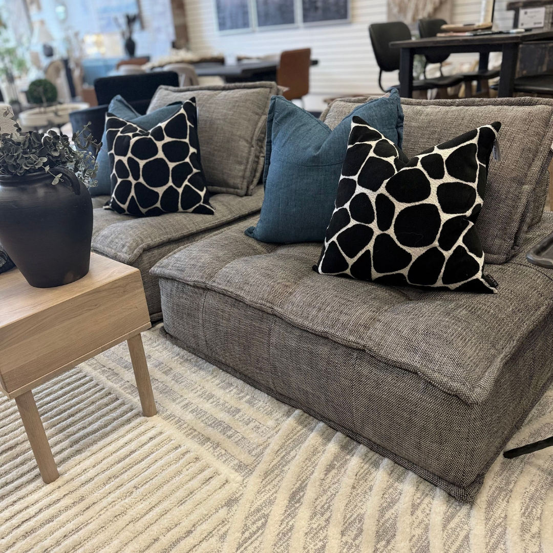 A cozy living room radiates modern elegance with a Princeton Sofa Lounger decked out in black and white patterned pillows and plush blue cushions. A small wooden side table supports a decorative black vase with plants, while a textured cream and gray rug covers the floor against blurred furnishings.