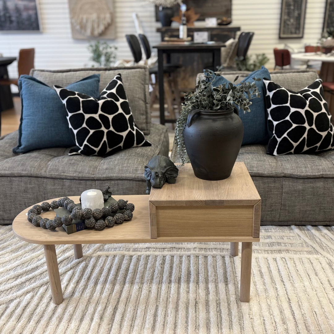 A sleek living room setup showcasing the Princeton Sofa Lounger features a wooden coffee table with a black vase of greenery, a decorative skull, and a round tray with a candle and beads. Plush cushions adorn the gray sofa over a textured white rug.