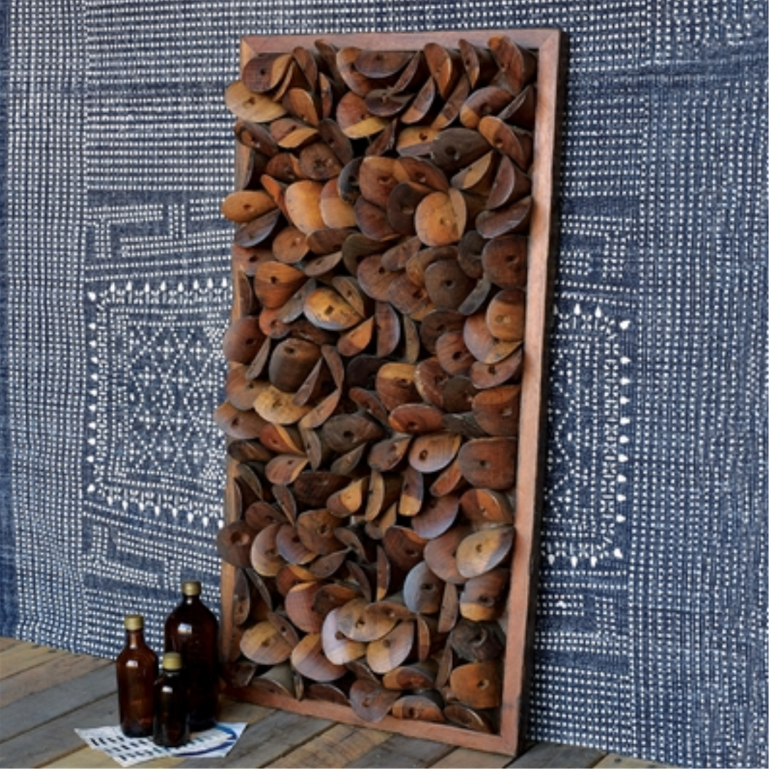 The Remarque Wood Wall Décor, crafted from reclaimed wood with overlapping rust-colored ovals, is displayed against a blue and white fabric. Nearby, three brown glass bottles and a stack of papers rest on the wooden floor.