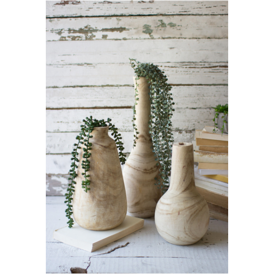 Three Cassidy Hand Carved Wooden Vases of different heights display cascading greenery and dried blooms, perfectly complemented by stacked aged books on a whitewashed wooden surface. Each distinct vase showcases its own shape against a backdrop of distressed white planks, adding a vintage charm.