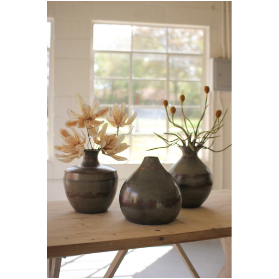 Three Finley Metal Vases with contemporary design sit on a wooden table in a sunlit room. The left vase holds beige flowers, the right one has stems with round seed heads, and the middle vase is empty. Large windows in the background let in natural light, creating a serene atmosphere.