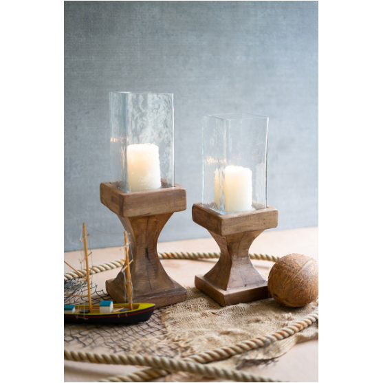Two lit candles in Mila Candleholders with glass covers sit on a table. Beside them are a decorative sailboat and coconut, all atop fishnet and rope. The neutral gray wall in the background enhances the rustic and cozy home decor atmosphere.