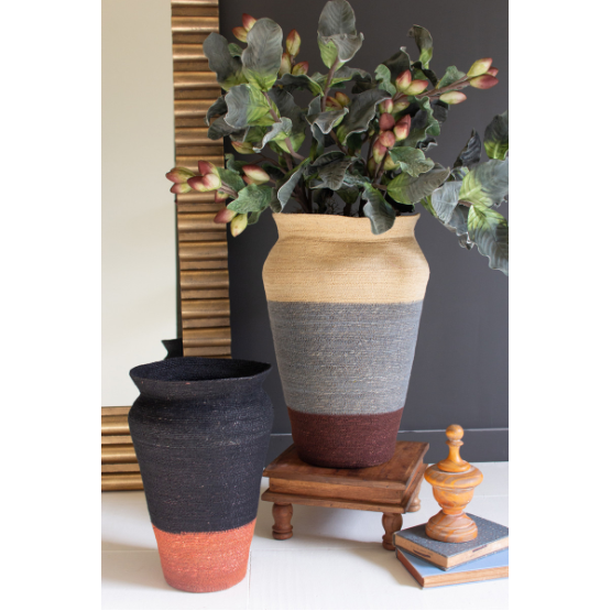 A Maren Rustic Seagrass Urn, featuring a gray and brown finish with green leafy branches, and a large black woven vase with an orange base sit on a small wooden platform. A striped wooden vase and two stacked books nearby enhance your home decor against a dark wall and a tall mirror with a striped frame.