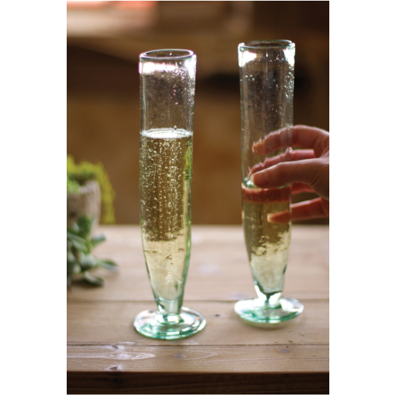 Two tall, slender Recycled Glass Champagne Flutes filled with bubbly champagne sit on a wooden table. A hand reaches for one glass as sunlight filters through a window, casting a warm glow. A small green plant is partially visible in the background, adding a touch of natural decor.