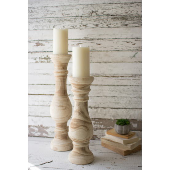 Two Huck Hand-Carved Wooden Candleholders, each supporting a cream-colored pillar candle, are elegantly displayed on a rustic white wood surface. Weathered white planks create a charming backdrop. To the right, a small stack of books and a tiny succulent in a gray pot complete this sophisticated statement piece.