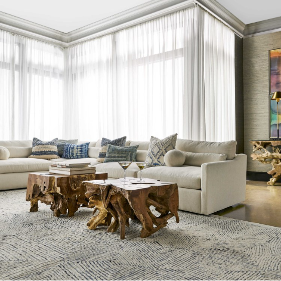 A modern living room features white sofas adorned with blue and patterned throw pillows, positioned beneath tall, sheer curtains. Two wooden Raiz Coffee Tables hold books and decor. The room has a light rug with a wavy pattern and colorful artwork on the wall.