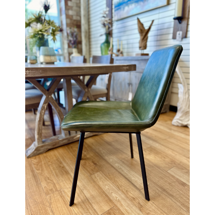 The Gabby Dining Chair features a seat and backrest upholstered in luxurious hunter green goat leather, supported by thin black iron frame legs. Positioned on a wooden floor, it sits next to a wooden dining table adorned with decor items. The background includes the Gabby Dining Chair, indoor plants, and a light-toned wall.