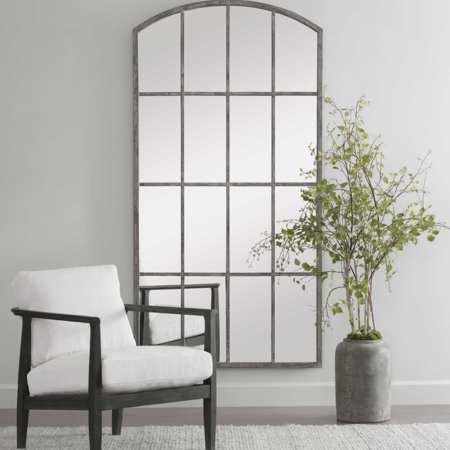 A white armchair with a black frame sits on a light gray floor beside a tall Potted River Birch Tree with green leaves, in front of an arched wall mirror that reflects soft light in a modern, minimalist room with pale gray walls.