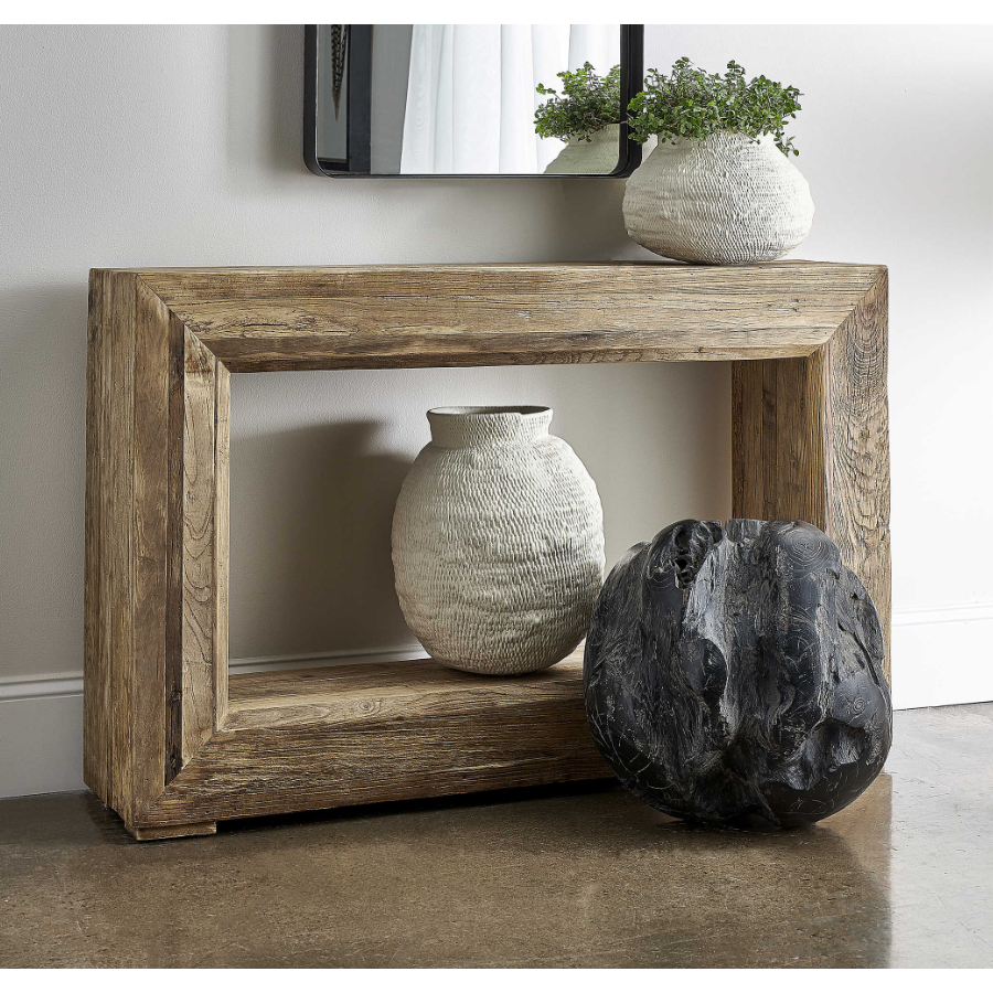 The Ridge Console Table, made from reclaimed elm wood, stands against a light grey wall. Textured white ceramic vases with green plants sit on and below it, and a mirror with a beveled open center design hangs above.