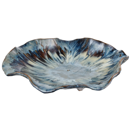 The Earthenware Charger is a handcrafted ceramic footed plate with an irregular, wavy edge reminiscent of a flower. It showcases a blue, brown, and cream reactive drop glaze that forms a swirling pattern. The glossy finish enhances the colors and textures, and its center is slightly concave.