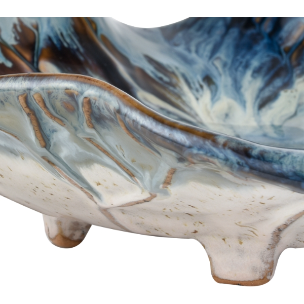 A close-up view of the Earthenware Charger reveals its decorative wavy edge. The reactive drop glaze creates a dynamic blend of blue, white, and brown tones that form a fluid, marbled design. The piece is elevated on small rounded feet, emphasizing its artisanal craftsmanship.