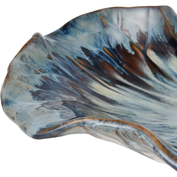 The Earthenware Charger showcases a close-up view of its wavy, scalloped edge, adorned with a swirling pattern in shades of blue, brown, and white that evokes the appearance of flowing water or a natural landscape. The reactive drop glaze imparts a smooth and glossy finish, enhancing the dynamic blend of colors.