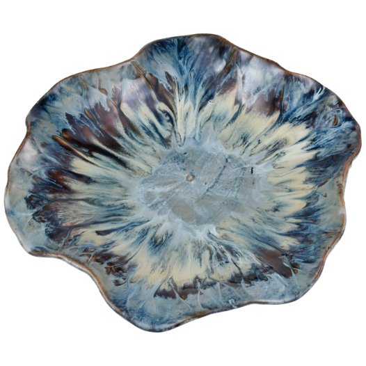 The Earthenware Charger is a ceramic plate with a footed base that showcases an irregular, wavy-edged rim. Its design features a marbled pattern in blue, cream, and hints of brown swirling outward like a floral motif. The reactive drop glaze provides a glossy finish that highlights the vibrant colors and organic shape of the plate.