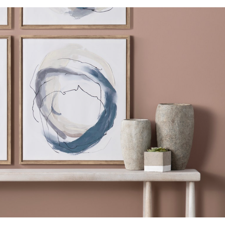 A minimalist setup with abstract art and vases. Two abstract paintings comprising circular strokes of blue, beige, and gray in wooden frames are mounted on a mauve wall. Below them is a slim console table holding two La Palata Vase in textured cement of different sizes and a small potted succulent, perfect for rustic home decor.